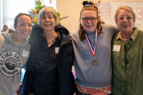 Four smiling women stand together indoors, one wearing a medal around her neck.