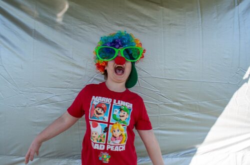 Child in a colorful wig and oversized glasses, wearing a Mario-themed shirt, makes a funny expression. Child in a colorful wig and oversized glasses, wearing a Mario-themed shirt, makes a funny expression.