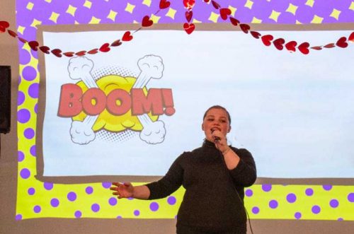 A person sings on stage with a BOOM! comic graphic and heart decorations in the background.